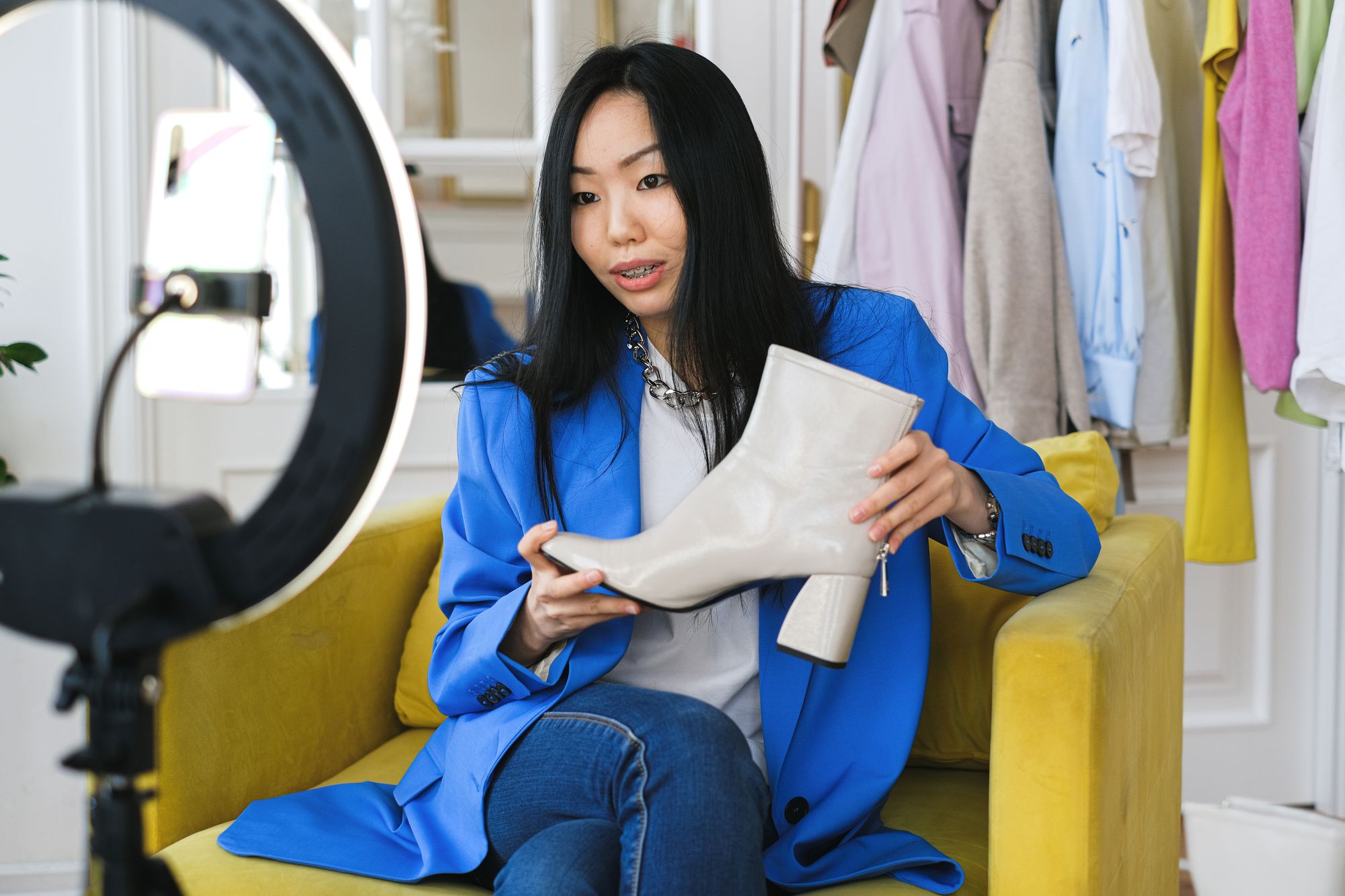 A live seller showing a white boot to her phone and ring light in front of a clothing rack